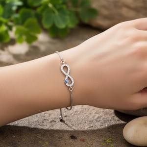 May include: Silver-toned bracelet featuring an infinity symbol adorned with clear crystals and a blue eye charm. The bracelet is delicate and adjustable, designed to be worn on the wrist. The background shows green foliage.