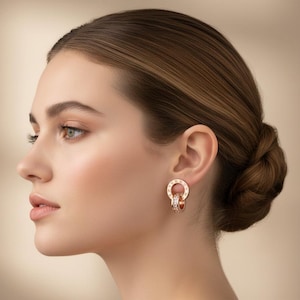 Puede incluir: Pendientes de oro rosa con un diseño circular y pequeños detalles brillantes. Los pendientes se lucen en la oreja de una persona, sobre un fondo suave y neutro. Los pendientes tienen un estilo clásico y elegante.