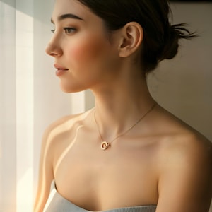 May include: A gold-colored necklace with two interlocking circles as a pendant. The necklace is delicate and sits on the person's collarbone. The person is wearing a light blue strapless top and is looking out a window.