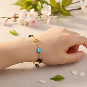 May include: A gold-colored chain bracelet featuring clover-shaped charms in various colors, including turquoise, yellow, and black. The bracelet is displayed on a person's wrist, with a soft background of flowers and leaves.