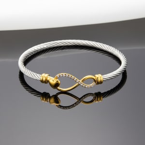 May include: A silver and gold-tone bracelet featuring an infinity symbol. The bracelet has a twisted cable design with gold accents and a gold infinity symbol adorned with small, clear crystals. A heart shape is on one end.