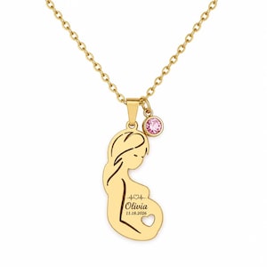May include: A gold-colored necklace featuring a pendant in the shape of a pregnant person. The pendant has the name "Olivia" and the date "11.18.2026" engraved on it, along with a heartbeat line and a heart cutout. A pink gemstone hangs from the chain.