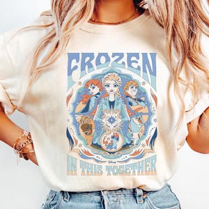 K&ouml;nnte beinhalten: Cremefarbenes T-Shirt mit einer farbenfrohen Grafik aus dem Disney-Film "Frozen". Das Design enth&auml;lt das Wort "FROZEN" in Blau und den Satz "IN THIS TOGETHER" in Hellorange. Die Grafik zeigt Charaktere aus dem Film.