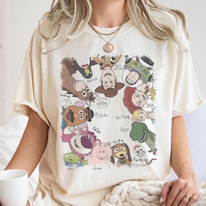 May include: Cream-colored t-shirt featuring a colorful graphic of various Toy Story characters, including Woody, Buzz Lightyear, and others. The names of the characters are written below them. The shirt has short sleeves and a relaxed fit.