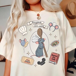 Può includere: T-shirt color crema con un'illustrazione a cartoni animati di uno chef ratto che tiene un cucchiaio di legno. Il design include il testo "Anyone can cook" e altre grafiche a tema culinario, come un cappello da chef e un libro di cucina.