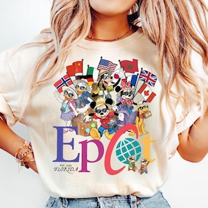 K&ouml;nnte beinhalten: Cremefarbenes T-Shirt mit einer farbenfrohen Grafik von Disney-Figuren, darunter Mickey und Minnie Mouse, Goofy, Donald und Daisy Duck sowie Pluto, die Flaggen aus aller Welt halten. Das Wort "Epcot" ist in Lila und Rot dargestellt, darunter "Est. 1982 Florida".