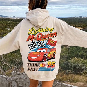 Può includere: Felpa con cappuccio beige con una grafica di Saetta McQueen del film Cars. Il design include il testo "Lightning McQueen" e "95", insieme a bandiere a scacchi e la frase "Think Fast".
