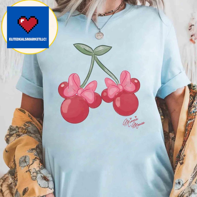 Op de afbeelding: Lichtblauw T-shirt met een cartoon kers ontwerp. De kersen zijn rood met roze strikken en groene stelen. De woorden "Minnie Mouse" staan eronder in cursief.