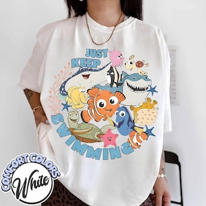 May include: White t-shirt with a colorful graphic featuring characters from the movie "Finding Nemo" and the phrase "Just Keep Swimming". The design includes fish, a turtle, and a starfish.