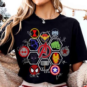 Pu&ograve; includere: T-shirt nero con una grafica colorata degli Avengers. Il design include vari loghi e firme di supereroi, con il logo centrale degli Avengers in rosso. La maglietta &egrave; a girocollo.