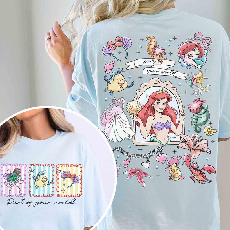 May include: Light blue t-shirt featuring a colorful graphic of Ariel from The Little Mermaid, seashells, and other related imagery. The text "part of your world" is also displayed on the shirt. The shirt has a crew neck and short sleeves.