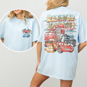 May include: A light blue t-shirt featuring a small graphic on the front left chest with a blue car and the text Name. The back displays a large, colorful illustration of Radiator Springs with Lightning McQueen and other car characters.