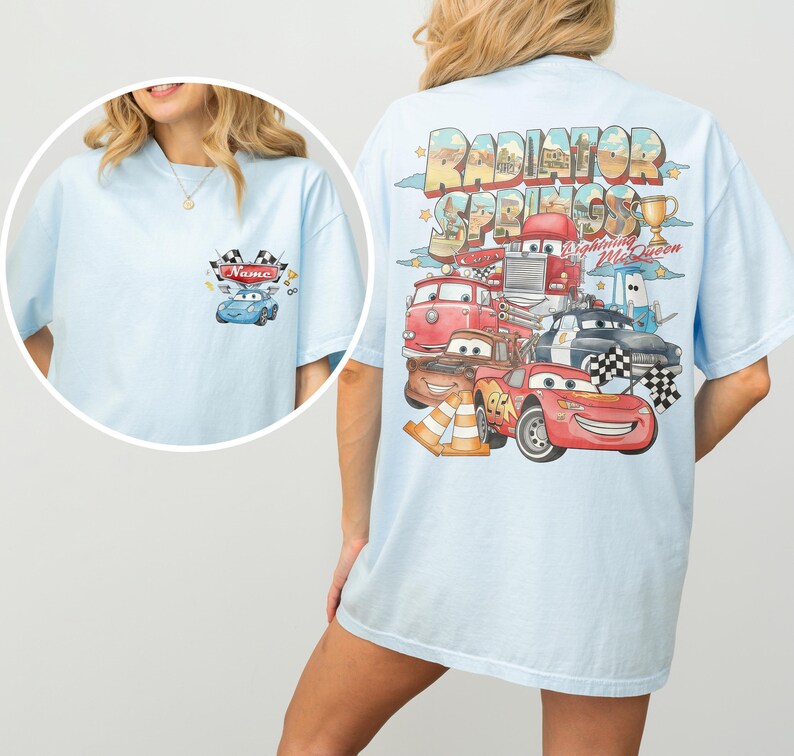 May include: A light blue t-shirt featuring a small graphic on the front left chest with a blue car and the text Name. The back displays a large, colorful illustration of Radiator Springs with Lightning McQueen and other car characters.