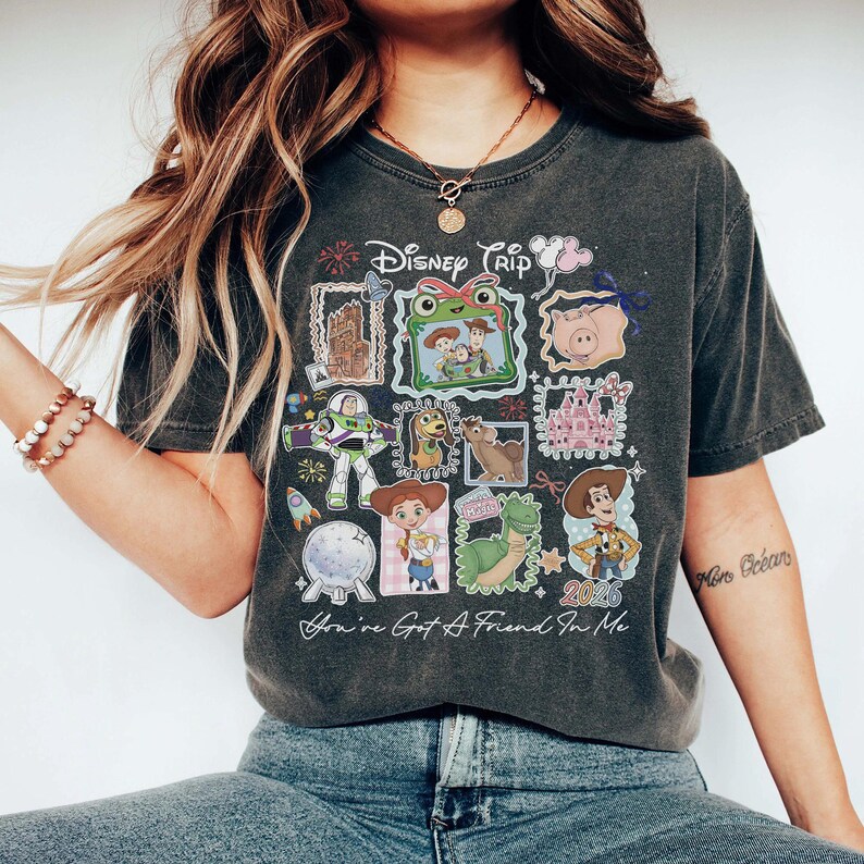 May include: A charcoal gray short-sleeve t-shirt featuring a graphic print with the text Disney Trip and You've Got A Friend In Me. The design includes various cartoon characters, a castle, and the year 2026 arranged in a collage of frames.