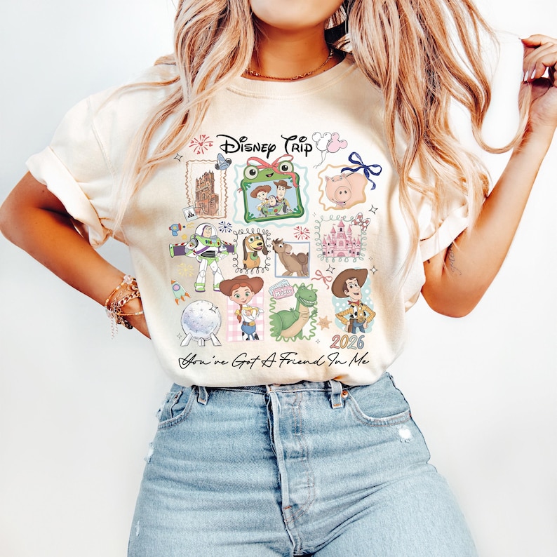 May include: A cream-colored t-shirt featuring a graphic print with the text Disney Trip and You've Got A Friend In Me. The design includes illustrations of various animated characters, a castle, and the year 2026 arranged in a collage style.