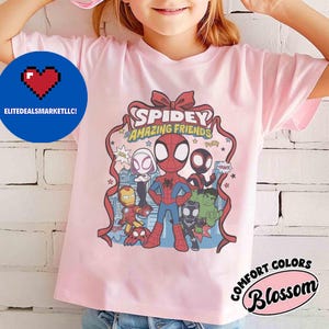 May include: A light pink t-shirt featuring a graphic of Spidey and his Amazing Friends. The design includes Spider-Man, Iron Man, Hulk, and other characters. The shirt also has the text "Spidey Amazing Friends".