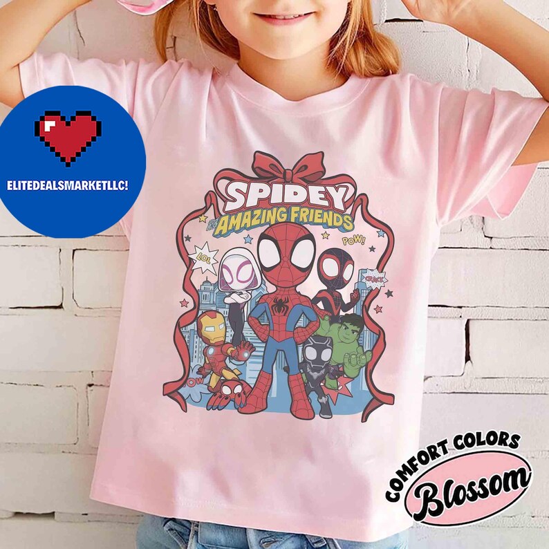 May include: A light pink t-shirt featuring a graphic of Spidey and his Amazing Friends. The design includes Spider-Man, Iron Man, Hulk, and other characters. The shirt also has the text "Spidey Amazing Friends".