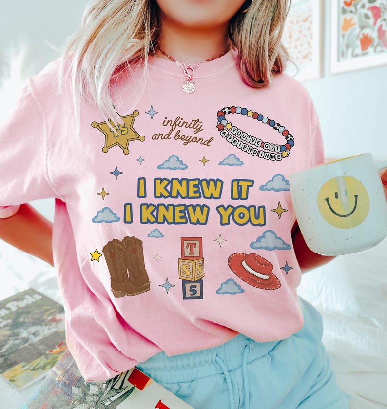 May include: A light pink t-shirt featuring the text I KNEW IT I KNEW YOU in yellow and blue. Graphics include a sheriff badge, a beaded bracelet with the text YOU'VE GOT A FRIEND IN ME, cowboy boots, alphabet blocks, and a red hat.