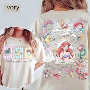 May include: Ivory-colored t-shirt featuring a colorful graphic design. The front displays three framed illustrations with the text "Part of your world." The back showcases a larger design with Ariel and other characters.