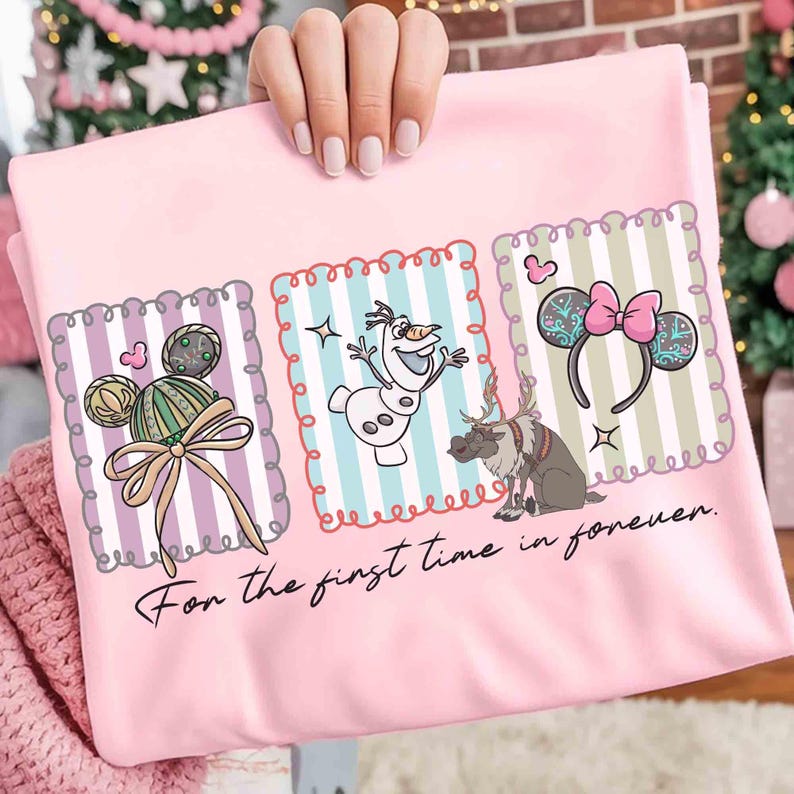 May include: Pink t-shirt featuring three framed illustrations of Disney characters. The illustrations include a Mickey Mouse-shaped ornament, Olaf the snowman, and reindeer. The text "For the first time in forever" is written in cursive.