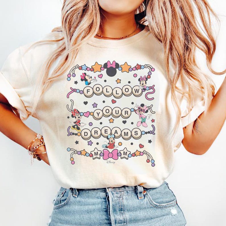 May include: Cream-colored t-shirt featuring a colorful graphic with the text "FOLLOW YOUR DREAMS" and various Disney character illustrations. The design includes Minnie Mouse, stars, hearts, and decorative elements.
