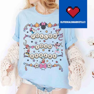 May include: Light blue t-shirt featuring the phrase "FOLLOW YOUR DREAMS" with Minnie Mouse characters and star accents. The shirt has rolled-up sleeves and a crew neck. The Disney logo is at the bottom.