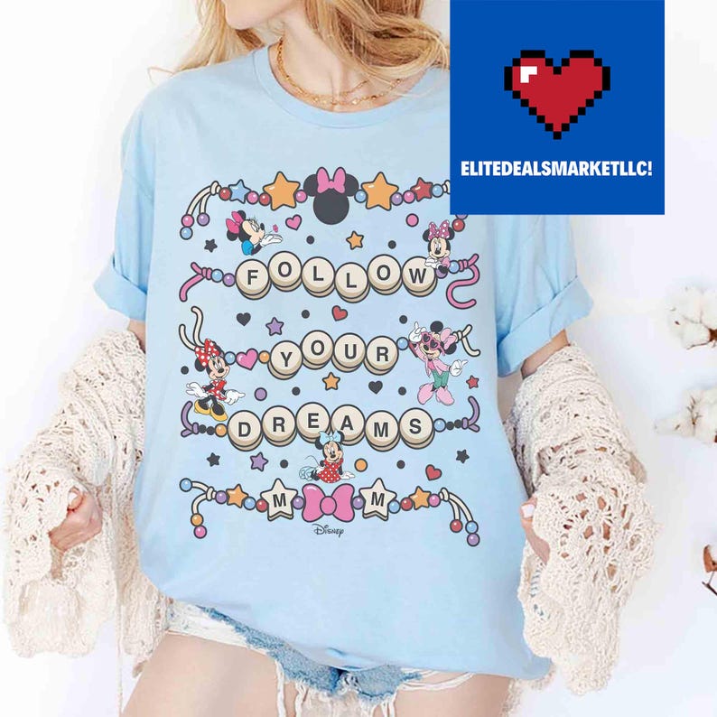May include: Light blue t-shirt featuring the phrase "FOLLOW YOUR DREAMS" with Minnie Mouse characters and star accents. The shirt has rolled-up sleeves and a crew neck. The Disney logo is at the bottom.