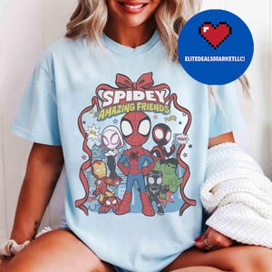 May include: Light blue t-shirt featuring a graphic of Spidey and his Amazing Friends. The design includes Spider-Man, Iron Man, Hulk, and other characters. The words "Spidey Amazing Friends" are displayed in a red ribbon design.