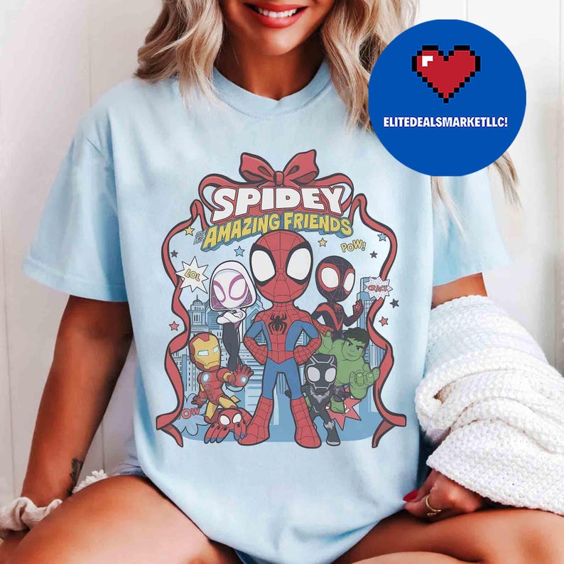 May include: Light blue t-shirt featuring a graphic of Spidey and his Amazing Friends. The design includes Spider-Man, Iron Man, Hulk, and other characters. The words "Spidey Amazing Friends" are displayed in a red ribbon design.