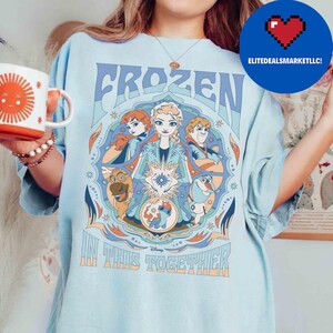 K&ouml;nnte beinhalten: Hellblaues T-Shirt mit dem Wort "FROZEN" in einer stilisierten Schriftart und Illustrationen von Charakteren aus dem Film. Der Satz "IN THIS TOGETHER" ist darunter gedruckt. Eine Tasse mit einem Sonnendesign wird in der linken Hand gehalten.