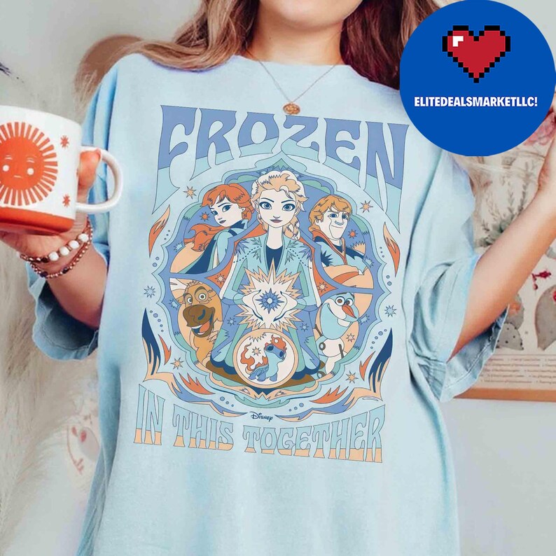 K&ouml;nnte beinhalten: Hellblaues T-Shirt mit dem Wort "FROZEN" in einer stilisierten Schriftart und Illustrationen von Charakteren aus dem Film. Der Satz "IN THIS TOGETHER" ist darunter gedruckt. Eine Tasse mit einem Sonnendesign wird in der linken Hand gehalten.