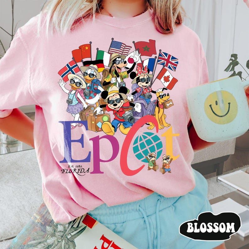 K&ouml;nnte beinhalten: Hellrosa T-Shirt mit einem farbenfrohen Design von Disney-Figuren, Flaggen aus aller Welt und dem Wort "EPCOT" mit "Florida" darunter. Das Wort "BLOSSOM" befindet sich in einem schwarzen Oval.