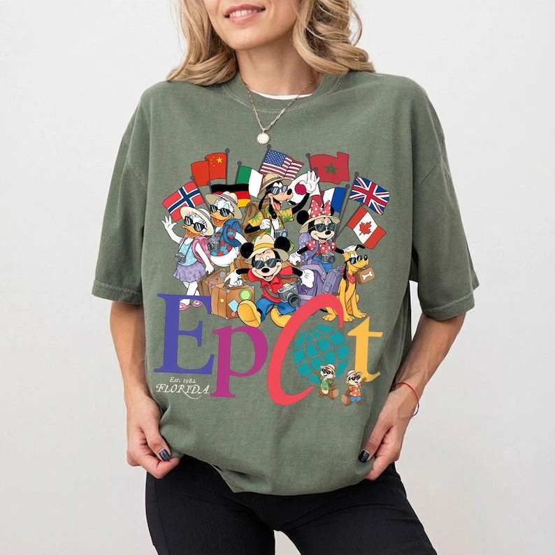 K&ouml;nnte beinhalten: Olivgr&uuml;nes T-Shirt mit einer farbenfrohen Grafik von Zeichentrickfiguren, darunter Micky und Minnie Maus, mit Flaggen aus aller Welt. Das Wort "EPCOT" wird in gro&szlig;en, farbigen Buchstaben angezeigt, darunter "Est. 1982 Florida".