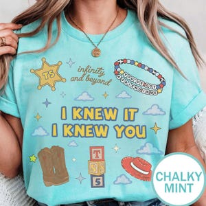 May include: A light mint green t-shirt featuring a graphic print with a yellow sheriff star, a beaded bracelet with text, brown cowboy boots, alphabet blocks, a red hat, and the phrases infinity and beyond and I KNEW IT I KNEW YOU with clouds.