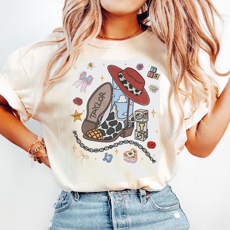 Puede incluir: Una camiseta color crema con un estampado gr&aacute;fico de una bota de vaquero con estampado de vaca con el nombre Taylor, un sombrero de vaquero rojo, bloques de alfabeto, un collar de cuentas con el texto IT THE WITY I K N E R Y O U y el n&uacute;mero 13.