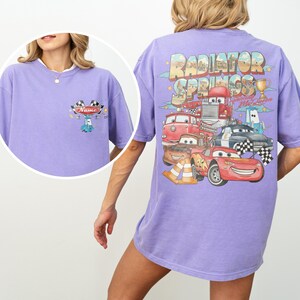 May include: A light purple t-shirt featuring a small graphic on the front chest with a blue car and checkered flags above the text Name. The back displays a large Radiator Springs graphic with Lightning McQueen, various characters, and a trophy.