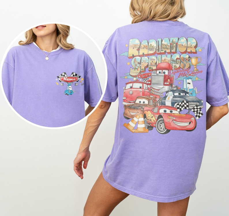 May include: A light purple t-shirt featuring a small graphic on the front chest with a blue car and checkered flags above the text Name. The back displays a large Radiator Springs graphic with Lightning McQueen, various characters, and a trophy.