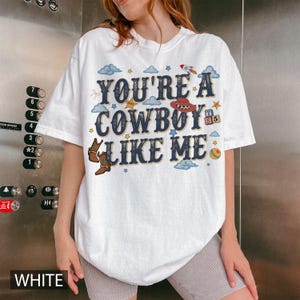 May include: A white t-shirt featuring the text "YOU'RE A COWBOY LIKE ME" in dark blue lettering. The design includes illustrations of brown cowboy boots, a red cowboy hat, a rocket ship, a colourful ball, small clouds, and yellow stars.