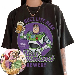 Może przedstawiać: Czarny t-shirt z grafiką Buzza Astrala trzymającego kufel piwa. Tekst głosi BUZZ LITE BEER, Star Command BREWERY oraz BEST Quality SINCE 1995. Okrągła wstawka przedstawia ilustrację trzech przytulających się postaci z animacji.