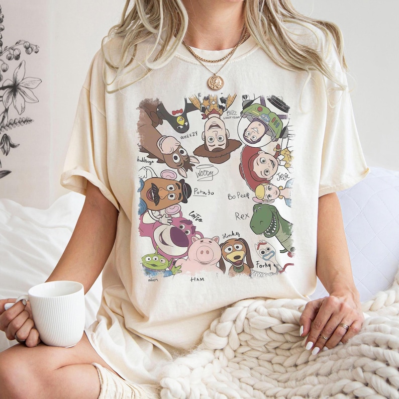 May include: Cream-colored t-shirt featuring a colorful graphic of various Toy Story characters, including Woody, Buzz Lightyear, and others. The names of the characters are written below them. The shirt has short sleeves and a relaxed fit.
