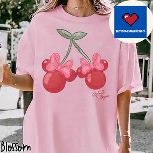 Op de afbeelding: Een lichtroze T-shirt met een cartoon kers ontwerp, Minnie Mouse strikken en de tekst "Minnie Mouse". Het shirt heeft ook de tekst "Blossom" rechtsonder en een blauw vierkant met een pixelhart en de tekst "ELITEDEALSMARKETLLC!"
