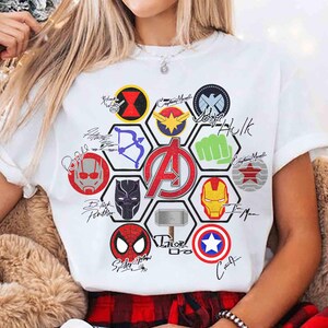 Pu&ograve; includere: T-shirt bianco con una grafica colorata raffigurante vari loghi e firme di supereroi, tra cui Iron Man, Captain Marvel e Spider-Man. Il logo degli Avengers &egrave; ben visibile al centro. La maglietta ha maniche corte.