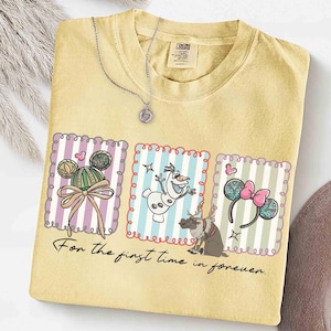 May include: Yellow t-shirt with three framed graphics featuring cartoon characters and accessories. The graphics include a Mickey Mouse-inspired design, Olaf the snowman, and Minnie Mouse ears. The text "For the first time in forever" is printed below the graphics. A silver necklace is draped over the shirt.