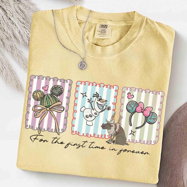 May include: Yellow t-shirt with three framed graphics featuring cartoon characters and accessories. The graphics include a Mickey Mouse-inspired design, Olaf the snowman, and Minnie Mouse ears. The text "For the first time in forever" is printed below the graphics. A silver necklace is draped over the shirt.