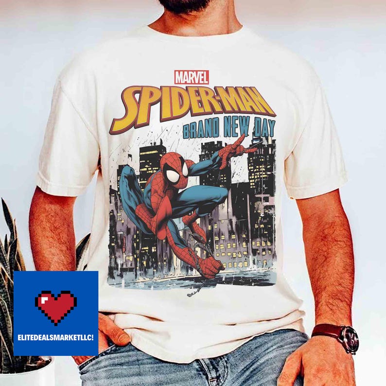 May include: Off-white t-shirt with a Spider-Man graphic, set against a city skyline. The design includes the Marvel logo and the words "SPIDER-MAN BRAND NEW DAY" in yellow and blue.