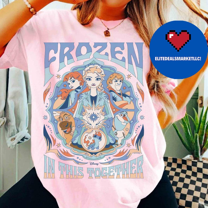 K&ouml;nnte beinhalten: Hellrosa T-Shirt mit dem Wort "FROZEN" in Blau und Bildern von Charakteren aus dem Film. Der Satz "IN THIS TOGETHER" ist darunter gedruckt. Das Design ist von einem dekorativen Rahmen mit Sternen und floralen Elementen umgeben.