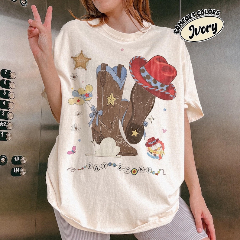 May include: An ivory t-shirt featuring a graphic print of brown cowboy boots, a red cowboy hat, a sheriff star, balloons, and the text TAY STORY. The design includes small stars, flowers, and a beaded string element across the bottom area.
