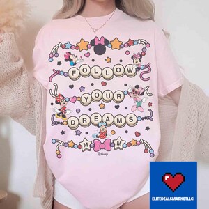 May include: Pink t-shirt featuring Minnie Mouse characters and the phrase "FOLLOW YOUR DREAMS" in a playful design. The shirt includes stars, hearts, and decorative elements, with the Disney logo at the bottom.