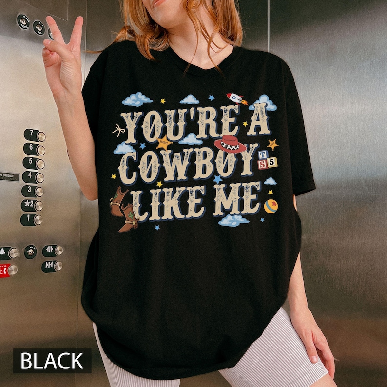 May include: A black short-sleeved t-shirt featuring the text "YOU'RE A COWBOY LIKE ME" in a distressed font. The graphic includes illustrations of a red cowboy hat, brown boots, a rocket, a yellow ball, and colourful alphabet blocks.