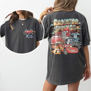 May include: A charcoal gray t-shirt featuring a small graphic on the front left chest with a car and the text Name. The back displays a large, colorful graphic of Radiator Springs characters including Lightning McQueen, with the text Radiator Springs.